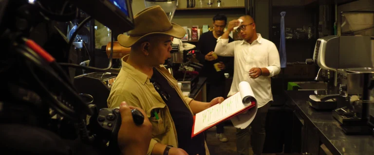 Man in a hat reading a script while the crew films in a kitchen