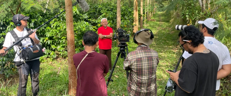 Film crew with camera and boom microphone filming between rows of coffee trees