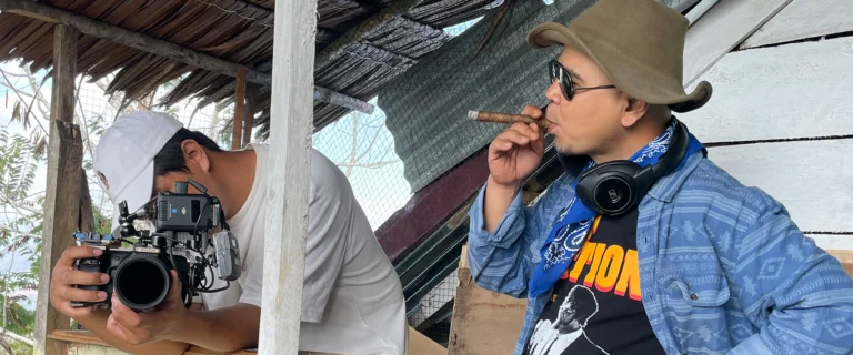 Cameraman beside a man in a hat and headphones smoking a cigar under a rustic roof