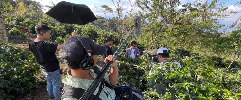 Crew filming on a coffee terrace, with an umbrella shading the camera and sound equipment