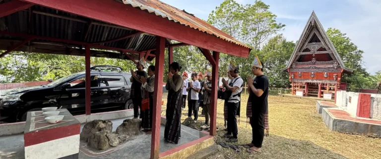 Crew and visitors gathered near a red-roofed pavilion and a traditional Batak house