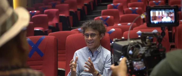 Interviewee speaking beside a film camera in a red-seated cinema hall