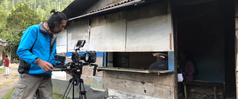 Cameraman in a blue jacket operating a camera on a tripod beside a small roadside stall