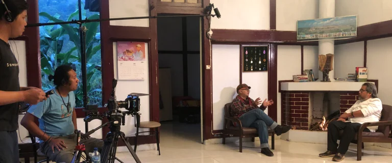 Crew filming an interview with two men seated in a traditional wooden house